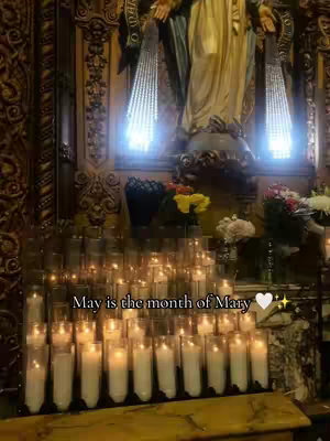 The image features a dimly lit, ornate interior, likely within a church setting. In the background, there's a statue of the Virgin Mary, partially visible, adorned in gold and blue. In the foreground, many lit candles are arranged, creating a warm glow. The text overlay 'May is the month of Mary' adds context and celebrates the focus of the video.