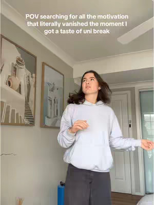 A young woman stands in a room, looking upward with a slightly dismayed expression. She wears a light blue hoodie and dark pants, and the room has light-colored walls with framed artwork. The lighting is natural.