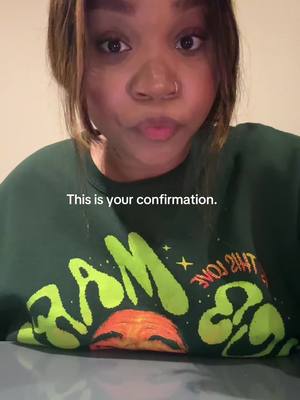 A selfie-style shot featuring a woman with dark hair and skin, looking directly at the camera with a slightly pursed expression. Overlayed on the image is the text "This is your confirmation." The woman is wearing a dark green sweatshirt with a graphic design in neon green and orange on the front, which is partially visible. The background is a neutral, blurred wall, keeping the focus on the woman.