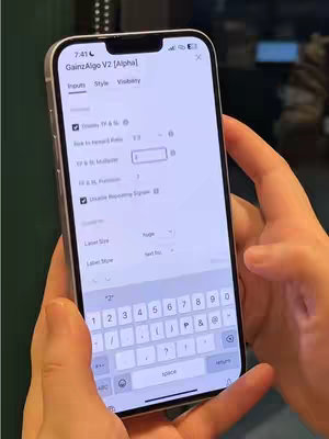 A person's hands holding a silver iPhone displaying the 'GainzAlgo V2 [Alpha]' app interface. The screen shows various input fields and settings with a visible keyboard at the bottom. The background is slightly blurred.