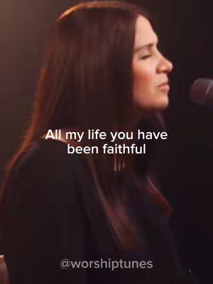 A woman with long, dark hair is seen singing into a microphone, with text overlaid on the image. The background fades from a dark tone on the bottom to a lighter tone on top. Text overlay is present, with the handle of the account at the bottom.