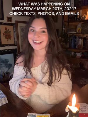 A woman smiling and looking at the camera, with overlaid text at the top. She is wearing a cream colored sweater and appears to be in a room with a wooden table in front of her. A lit candle is on the table,  with some tarot cards subtly visible. There is a warm, soft lighting.