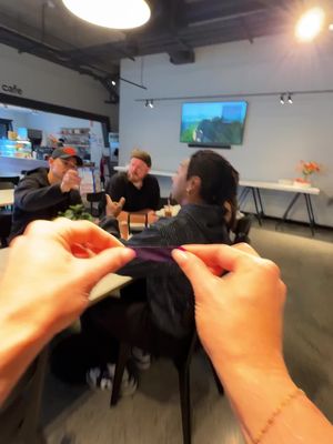 The image shows a first-person perspective in a cafe. A person's hands are holding a purple object, possibly a strip or a piece of something, in the foreground. In the background, there are several people sitting at a table, engaged in a conversation. The lighting is bright, and the setting appears casual and social.