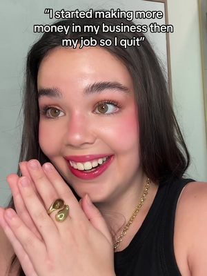 A selfie-style shot of a woman with vibrant makeup and a gold ring. She is smiling with her hands near her face. The background appears to be plain or slightly out of focus.