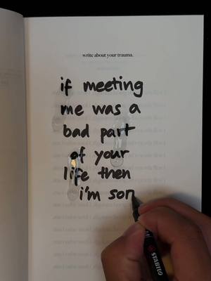 An open book is displayed with text written in black marker towards the center of the page. The text reads: 'write about your trauma.  if meeting me was a bad part of your life then i'm son[missing word]'.  A hand holding a black pen is situated at the bottom right side of the page. The background is a dark gray.