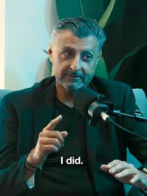 Image shows a man with silver hair, wearing a dark jacket over a black shirt, sitting in an armchair. He’s holding and speaking into a microphone with a focused expression, pointing with his index finger. Behind him are blurred green and blue elements and a plant.