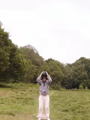 A man in a patterned jacket and white pants stands on a grassy path with his hands raised near his head. The background has a forest and an overcast sky