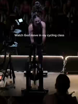 A woman is riding a stationary bike in a dimly lit room. She is wearing a sleeveless top and is smiling. There are other people on stationary bikes in the background. Text is overlayed.