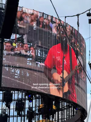 A live concert scene is captured from a low angle, featuring a large screen displaying a performance. The screen shows a person holding a guitar on stage, framed by a background of an audience. Below the performer, the caption 'best opening act everrr at ed sheeran's concert' is written.