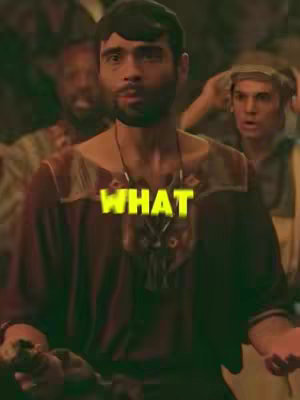 A somewhat grainy, medium shot from a historical drama, featuring a man in a maroon tunic with gold accents on a brown background. The man has a surprised expression with the word "WHAT" in a yellow, bold font across his chest. Other people in the background have expressions of shock and curiosity