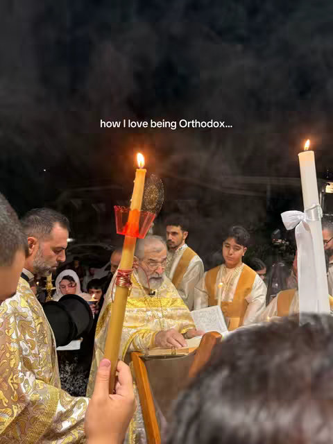 A nighttime scene depicts a religious ceremony with several people, including a priest holding a candle. The background is dark, and the lighting primarily comes from candles. The composition is vertical, likely taken with a phone.