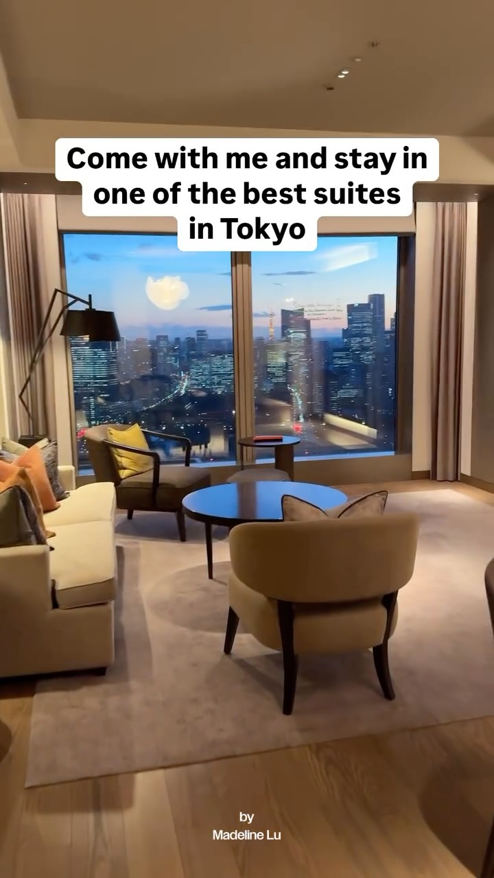 The cover image shows a brightly lit, luxurious suite in Tokyo. A large window dominates the background, offering a stunning view of the city skyline at dusk, with buildings and city lights visible. In the foreground is the suite's interior, featuring a cream-colored sofa, two armchairs, a round coffee table, and a floor lamp. The room is decorated in neutral tones with an emphasis on natural light and modern design. The image is well-composed, creating an inviting and elegant atmosphere.