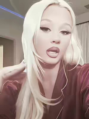 A close-up shot of a woman with long, platinum blonde hair. She is wearing a dark, silky top and has full lips and a direct gaze. Some background elements indicate a bedroom setting.