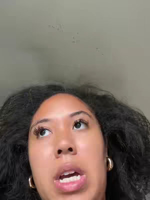 A close-up shot of a woman's face looking upwards. Her dark, curly hair frames her face. She is wearing gold hoop earrings, and her mouth is slightly open as if speaking. The background is a blurred, neutral color. The lighting appears to be soft, highlighting her facial features.