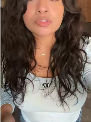 A selfie-style shot with a woman in the foreground; her head fills most of the frame. She has dark, wavy hair, and a pale complexion. She wears a light-colored, possibly white, long-sleeved shirt. Visible text overlay at the bottom says "SO II". The background is blurred, suggesting an interior setting. The woman is looking slightly upward.