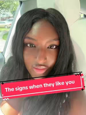 A selfie-style video featuring a person with long, dark hair. The person is situated inside a car, as suggested by the visible portion of a car seat and exterior views through the window. The main subject is a young woman with a nose piercing. The background outside the car includes a view of grass and trees, suggesting a daytime setting. A red banner with text is overlaid on the image.