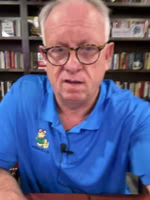 A middle-aged man with glasses and a blue polo shirt speaks directly to the camera against a backdrop of bookshelves.