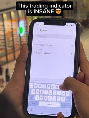 A person's hands holding a smartphone, displaying the TradingView app with a search bar filled with the text "Gainzalgo" and the "GainzAlgo V2 Alpha" indicator is shown. A text overlay at the top reads, "This trading indicator is INSANE".