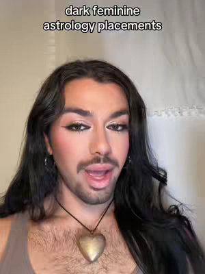 The cover image is a selfie featuring a person with long dark hair and significant makeup. The text overlay reads "dark feminine astrology placements." The person is wearing makeup, including dark eyeliner, blush, and lip color. They are wearing a gray top and a necklace with a heart pendant. The background appears to be a neutral wall, and the overall aesthetic is stylized with a focus on the person's face.
