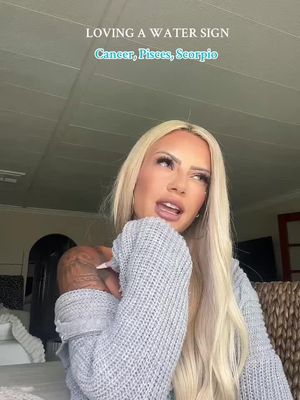 The image shows a fair-skinned woman with long, light blonde hair. She is wearing a gray sweater and has a tattoo visible on her left shoulder. Written text overlays appear at the top of the screen, and the background shows portions of a room with lighting fixtures and a textured surface on the right side. The woman is mid-speech, leaning into a hand-on-chin gesture. The overall tone seems informational or opinion-based.