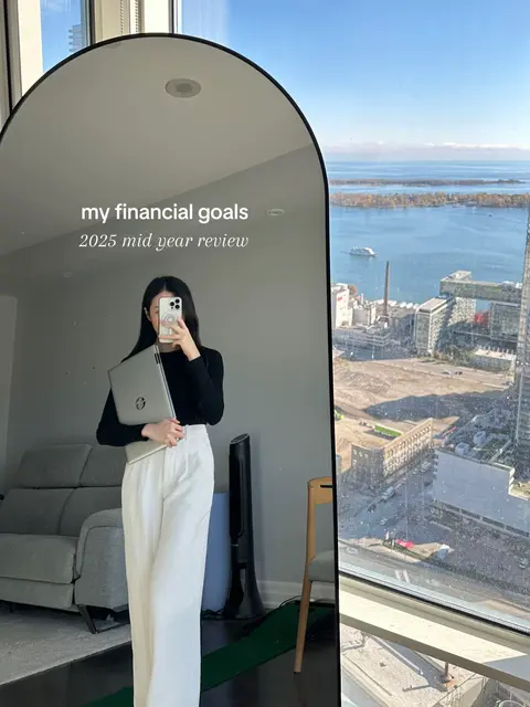 A woman stands in front of a large arched mirror holding a laptop and her phone, taking a selfie. The mirror reflects a cityscape. The woman is wearing a black top and white pants.