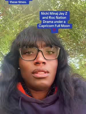 A selfie of a woman with dark hair, bangs, and large black-framed glasses. The background is blurred with green foliage, suggesting an outdoor setting. Overlay text in a blue box provides context regarding Nicki Minaj and ongoing dramas.