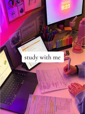 A desk lit with pink and purple ambient lighting. The scene includes a laptop, a tablet, papers, a pen, and a computer monitor displaying 9:23 in a glowing font. Highlighters, pens, and decorative items add to the study environment. One hand is visibly writing on a paper. The text overlay says 'study with me'.