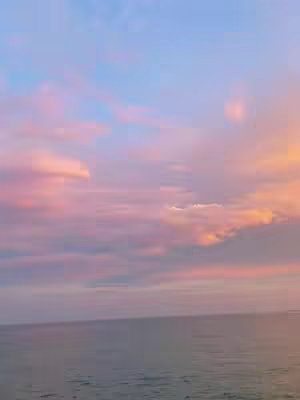 A scenic shot of a body of water under a colorful sky. The sky transitions from a vibrant light blue to soft pinks and oranges, with wispy clouds scattered throughout. The water has gentle ripples, reflecting the sky's colors. The overall composition evokes a sense of peace and tranquility.
