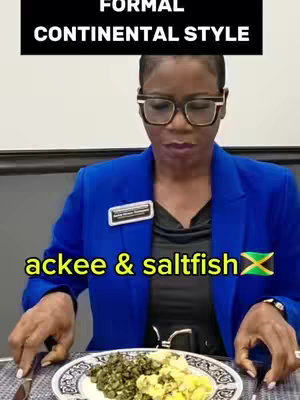 The cover image shows a woman seated at a table, preparing to eat a meal. She wears eyeglasses, and a blue blazer with a name tag. The dish in front of her is white with a black patterned edge, containing a portion of green leafy vegetables and what one might assume to be a portion of yellow rice. The bottom of the image has a yellow banner with the text "ackee & saltfish", along with the Jamaican flag.