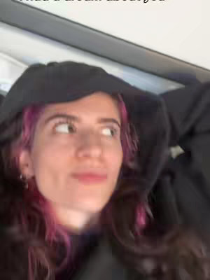 A woman is looking to her left against an out-of-focus background. She is wearing a black hoodie. Her hair is styled with pink highlights. The overall lighting is soft.