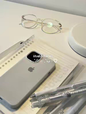 A well-lit, top-down composition presents a collection of study-related items. A pair of clear-framed glasses sits on top, next to a partially visible notebook. Below, a gray phone case displaying the Apple logo is centered. A transparent binder, several pens, and lined paper are also visible, creating a neat and organized aesthetic.