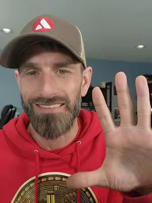 A man in a red hoodie and a baseball cap is holding up his right hand with fingers spread, indicating the number five. He is smiling at the camera. The background is blurry and appears to be an indoor setting.