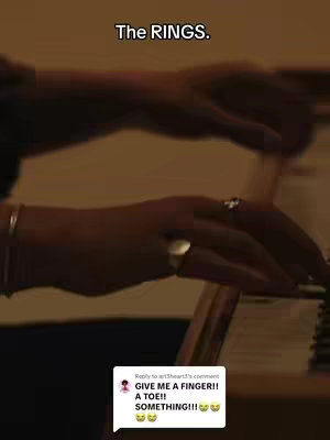 A close-up shot highlights a person's hand, emphasizing the rings they are wearing. The hand is positioned with rings on the fingers and a gold watch on. The background is dimly lit, allowing the rings and the hand to stand out.
