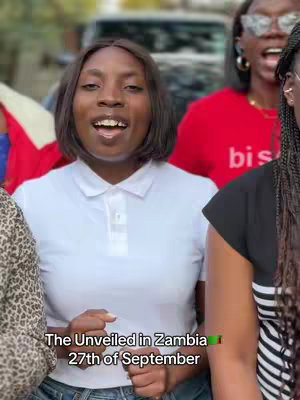 A group of four women are standing outdoors with a vehicle visible in the background. The central figure, likely the content creator, is a black woman in a white polo shirt. Text overlayed reads, "The Unveiled in Zambia 27th of September."