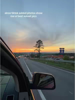 View from inside a car driving on a highway at sunset. The sky transitions from light blue to orange and yellow at the horizon. There is a lone tree in the middleground and signs in the background.