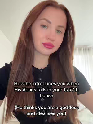 A woman with long, auburn hair and light skin is centered in the frame. She has bright pink lipstick and a small, subtle smile. Behind her, a neutral room setting includes a white wall and a portion of a window covered by a curtain. A vertical block of text is placed below the woman. The composition is simple and well-lit.