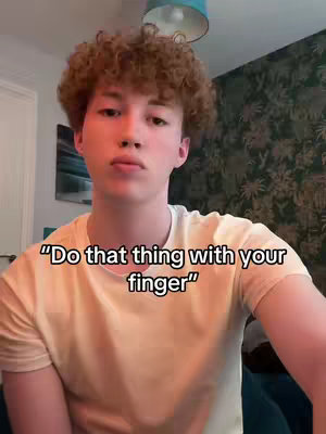 A young man with curly brown hair is the focus of the shot, with the foreground featuring his head and shoulders. He's wearing a light yellow t-shirt and appears to be indoors, with a colorful patterned wall and a lamp visible in the background. Text overlayed on the image says, "Do that thing with your finger."