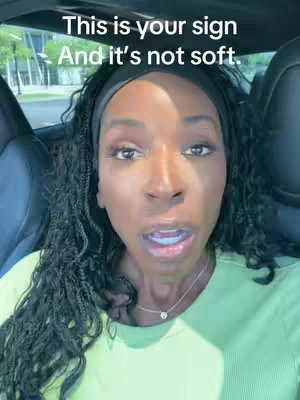 A woman with locs and a headband is seated inside a car, looking towards the camera with a slight open-mouth expression. The upper portion of the car’s interior is visible.