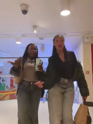 The cover image shows two young women standing in a brightly lit hallway of a shopping mall-like building. The woman on left is of darker skin and is wearing a brown top and jeans. She is holding an insulated cup in her left hand and gesturing with her right hand toward the right. The woman on the right is fair-skinned, wears a black top and jeans, and is also gesturing towards the right side of the image while wearing a surprised expression. They appear to be reacting to something off-camera. The background includes the ceiling, lights, and a security camera.