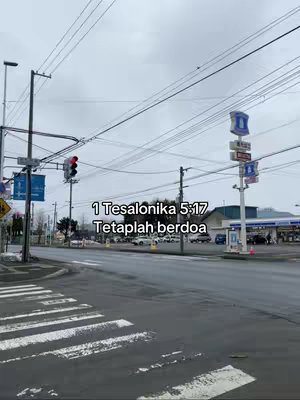 A street scene on a cloudy day. The shot focuses on a road with a crosswalk, leading towards a distant building. Power lines clutter the sky. Text in Indonesian is overlaid.