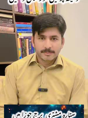 A man in a collared shirt with a small microphone is seated. A shelf filled with books is in the background. There is a graphic at the bottom with text in a different language.