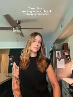A woman stands indoors in what appears to be a kitchen or living area, facing the camera. She has long, wavy blonde hair and is wearing a black top with visible tattoos on her arm. The background shows elements of a home setting, including a ceiling fan, cabinets, and pictures on the refrigerator. The overall composition is lit by natural light and the framing emphasizes the woman's upper body and the surrounding environment. The text overlay is placed at the top, centered.