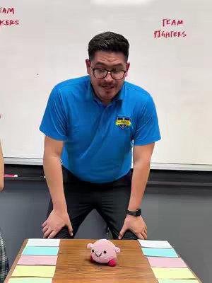 A man in a blue polo shirt and glasses stands behind a table with a colorful placemat and a small plush toy. Behind him is a whiteboard with text.