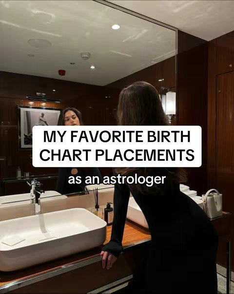 A woman in a black dress stands in a modern bathroom, looking into a large mirror. The bathroom has dark wood paneling and a white sink. A text overlay in a white box states "MY FAVORITE BIRTH CHART PLACEMENTS as an astrologer."