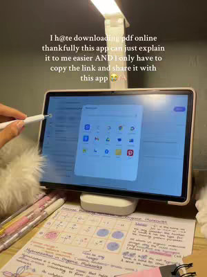 A hand is holding a stylus, pointing at a tablet screen, highlighting an app icon. The screen displays colorful icons. The table is set against a backdrop of study notes and a desk lamp, enhancing the setting.