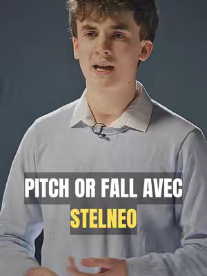 A young man with light skin is centered in the frame, looking slightly to the side while talking. He is wearing a light blue sweater with a white collared shirt underneath. A small microphone is clipped to his shirt collar. The background is a plain, dark gray. Text overlays appear at the bottom, with a dark gray background, and white and yellow text.