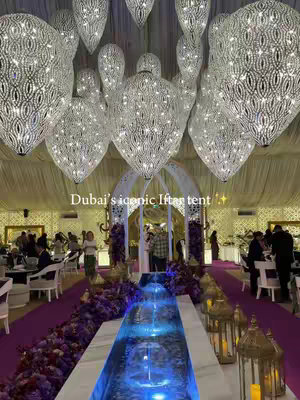 An interior shot of a tent decorated for an event, likely iftar. Large, ornate chandeliers hang from the ceiling, reflecting light. A central water feature with blue lighting runs along the floor, flanked by purple floral arrangements. Tables and chairs are set up, and people are visible, suggesting a dining area. Gold accents and warm lighting add to the elegance.