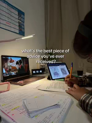 A desk setup with a laptop displaying a video and a tablet with notes, a calendar on the wall and a desk lamp.
