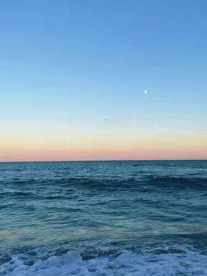 A serene seascape at dawn or dusk. The sky transitions from a soft blue at the top to a warm peach and pink horizon. The ocean has gentle waves. The foreground is filled with the foamy surf breaking on the shore. The top center of the image has the moon.
