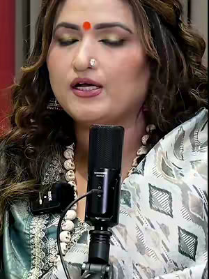 A medium shot shows a woman with long, wavy brown hair and elaborate makeup, including prominent eyeshadow, wearing an ornate saree. She's positioned against a blurred background, and has visible accessories. A microphone is positioned in front of her face.